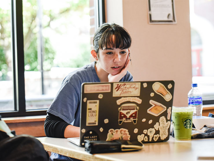 A young adult looks at an open laptop, chin in hand.