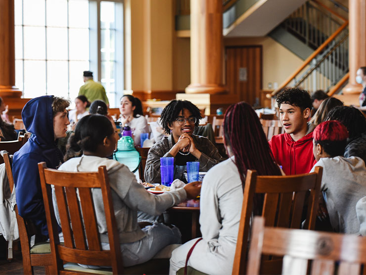 A large group of students seated around a table chat and laugh at a dining hall.