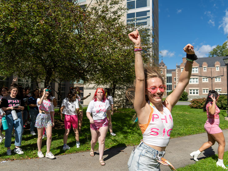 A college student with pink, heart-shaped sun glasses runs, arms extended, while others cheer nearby.