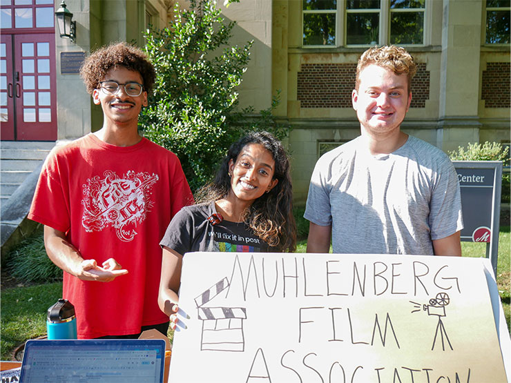 Three young adults smile at the camera while outdoors, pointing at a handmade sign that reads 