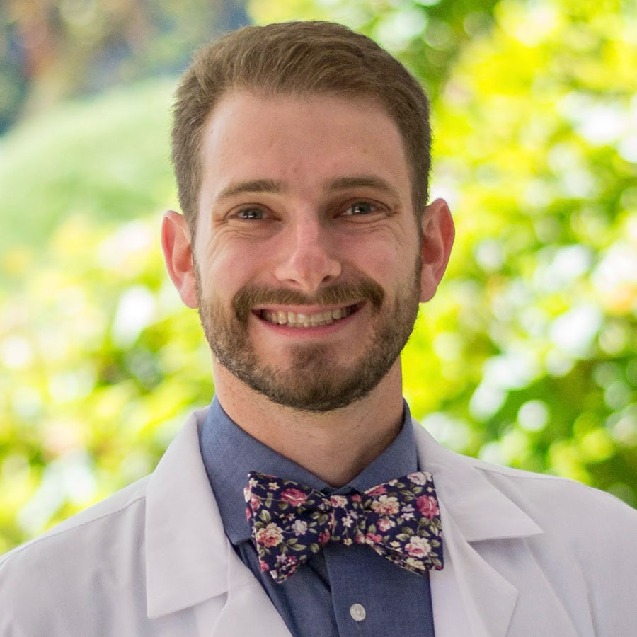 Man wearing white jacket and blue collared shirt with bowtie standing outdoors in front of green foliage.