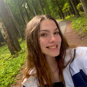 A college student with long brown hair smiles at the camera in a selfie taken along a forested path.