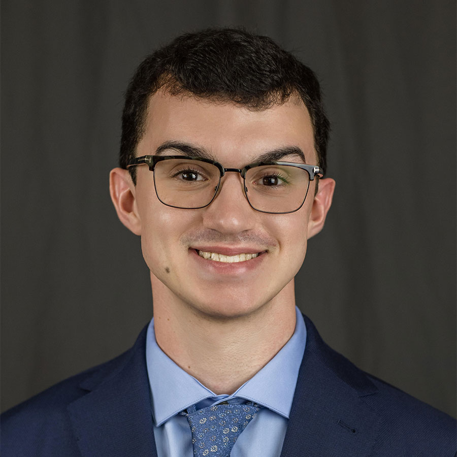 A young man wearing a suit and glasses smiles at the camera.