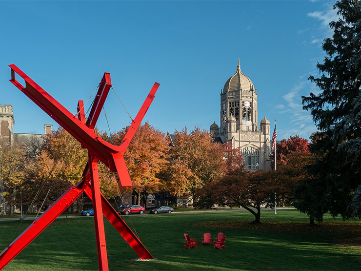 The bright red, modern sculpture stands in the foreground with the Haas College Center clocktower in the distance on a sunny fall day on the campus of Muhlenberg College.