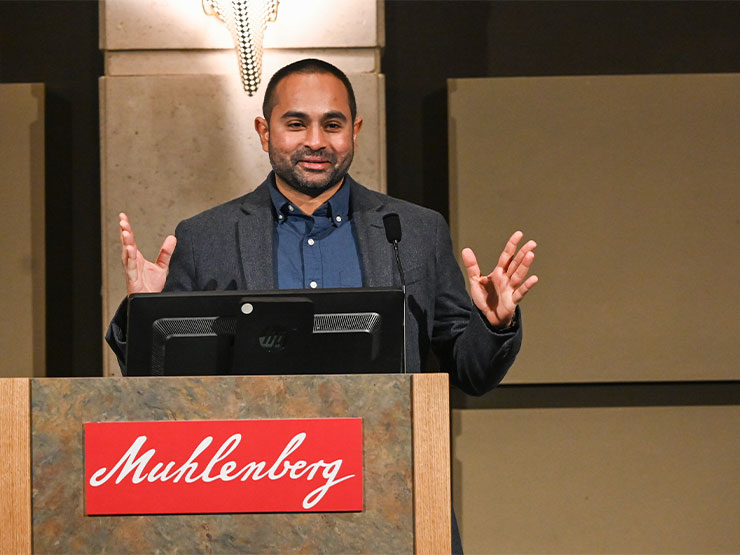 An adult in a dark grey suit with a dark blue shirt speaks at a podium labeled 