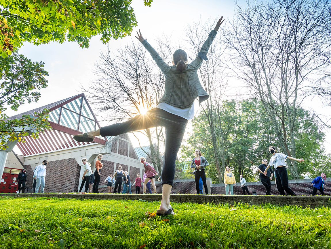 A college student raises their arms in a triumphant pose on a lawn in front of other college students and a library