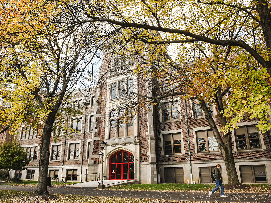 A college student walks past an academic building and trees with colorful fall foliage