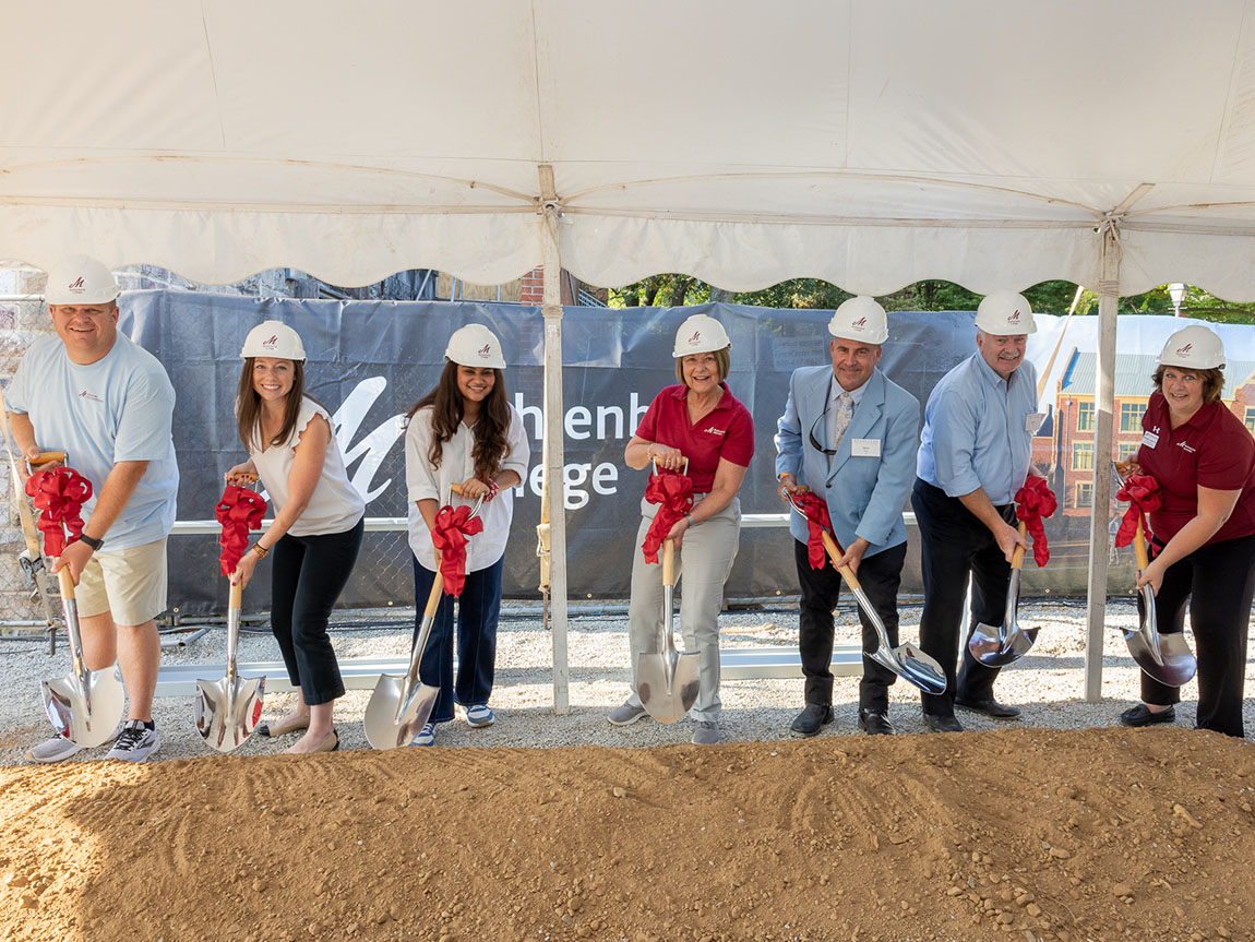 A group of seven people wearing white hard hats pose for a photo holding shovels with red bows on them