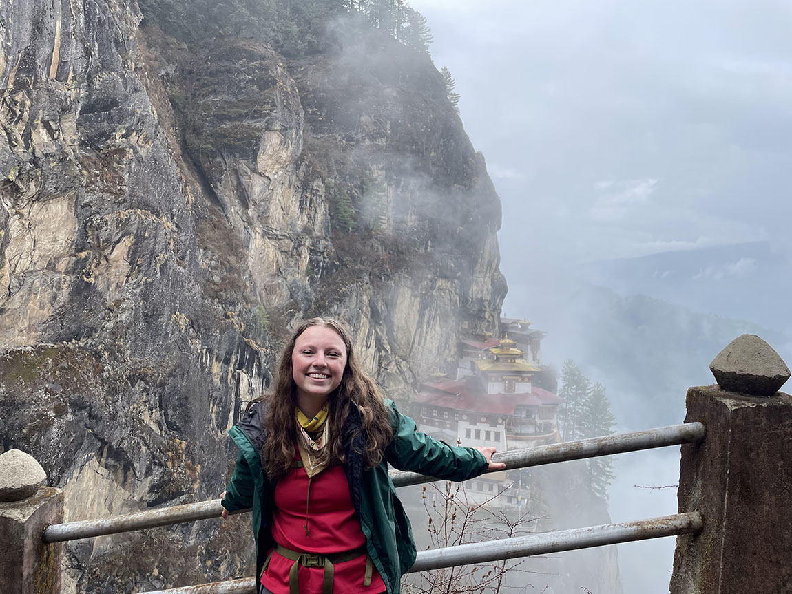 A smiling college student leans against a railing with a building perched on the side of a mountain visible behind fog in the background