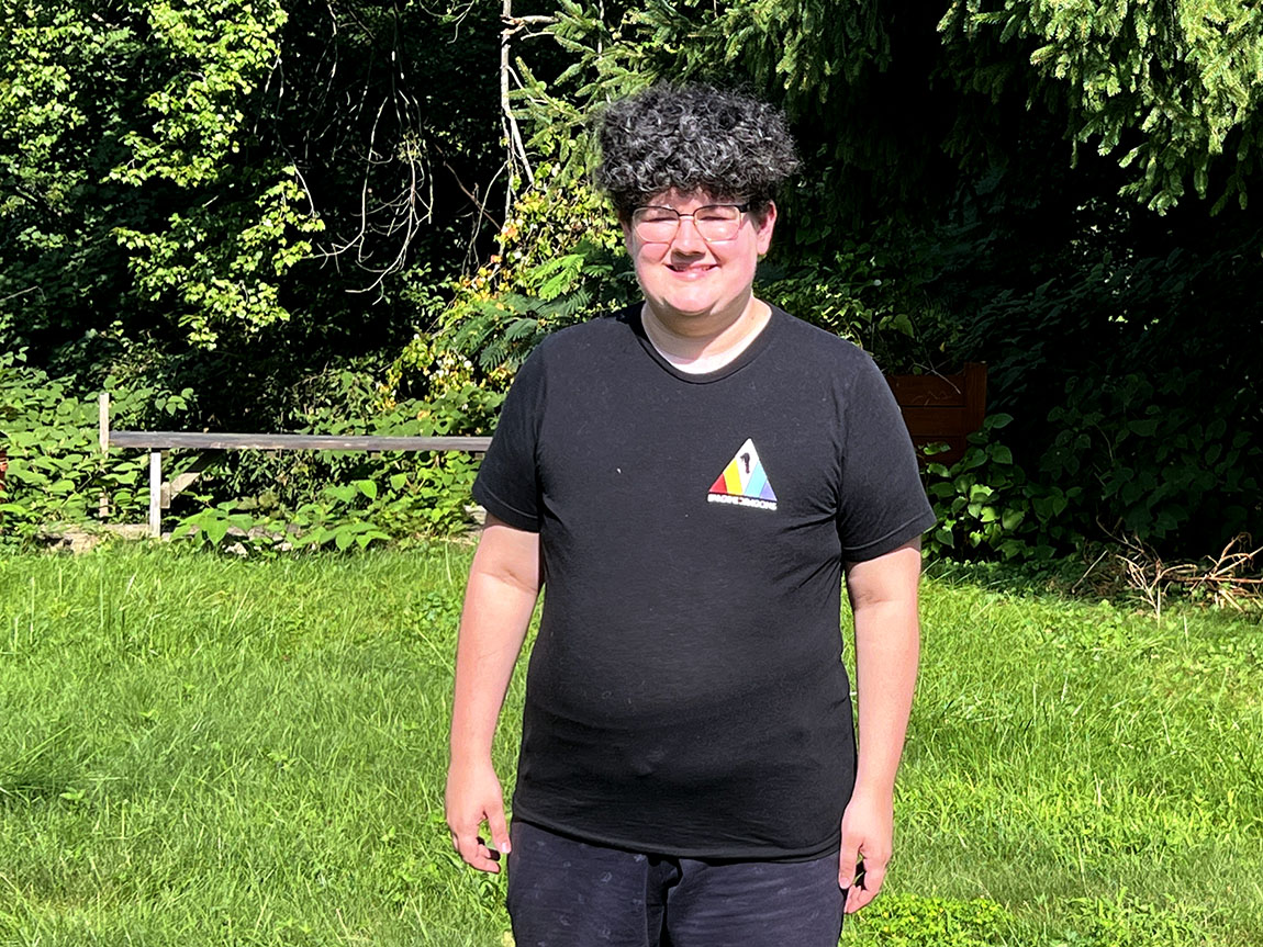 A college student wearing a black shirt and black shorts smiles for a portrait outside