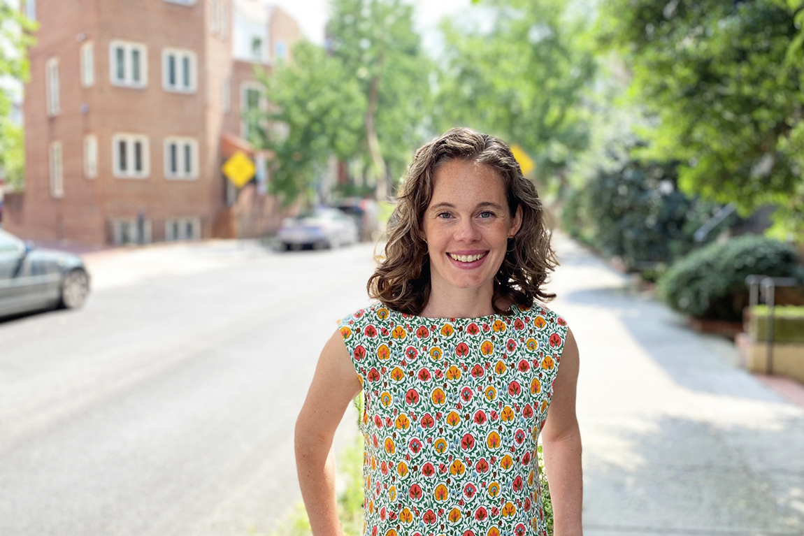 A white woman with curly brown hair wearing a floral dress stands on a city street