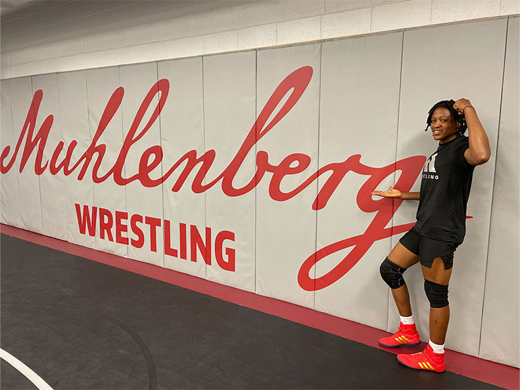 A college women's wrestling coach stands in front of a gym wall that says Muhlenberg WRESTLING