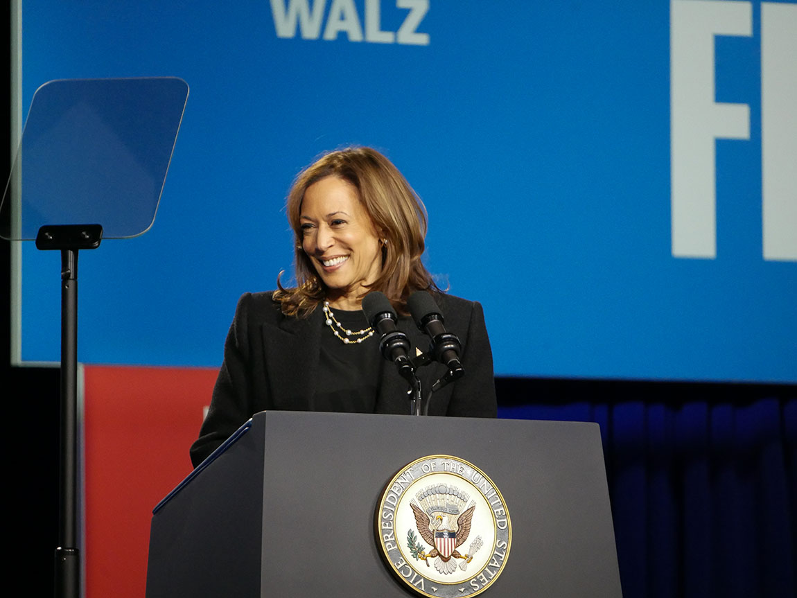 A woman stands at a podium with the vice presidential seal on it and smiles