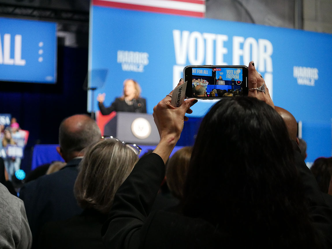 A rally attendee takes video on a cell phone of a woman speaking at a podium on stage