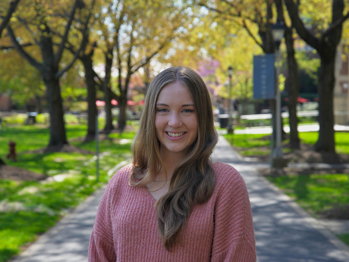A college student in a pink sweater stands on academic row