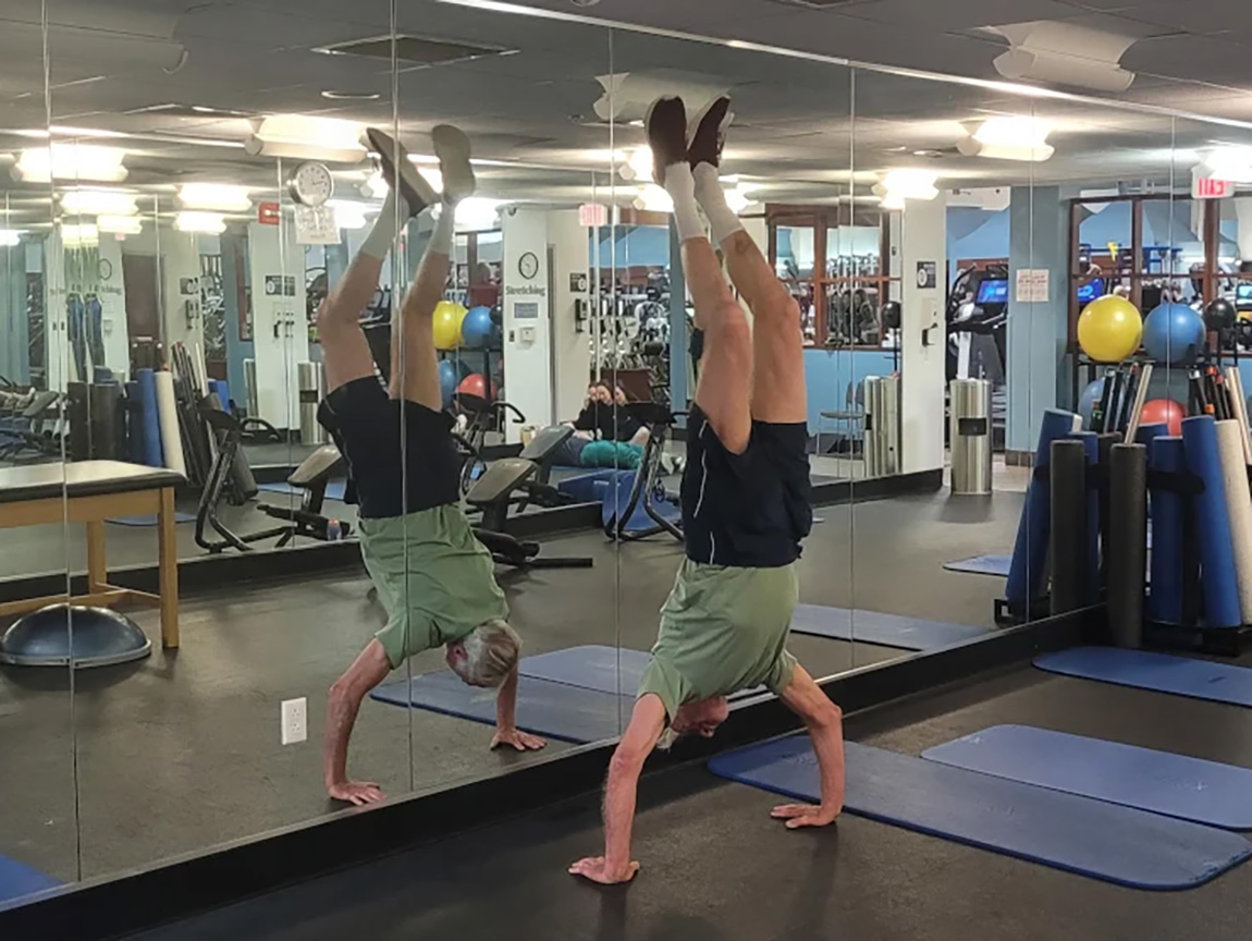 An older man does a handstand in gym clothes in front of a full-length mirror