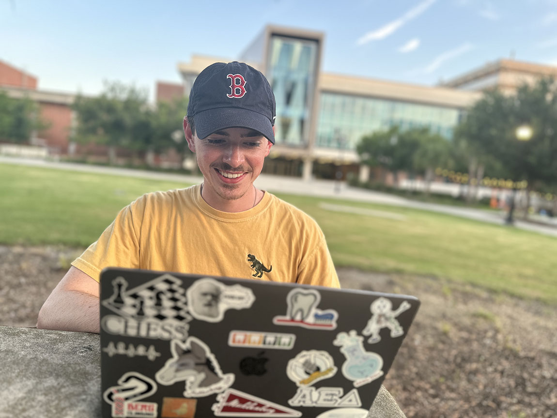 A college student wearing a Red Sox hat smiles while working on his laptop outside