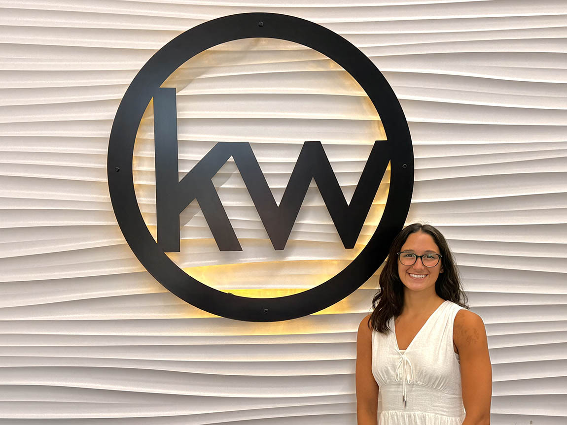 A college student stands next to a big circular sign with the letters 