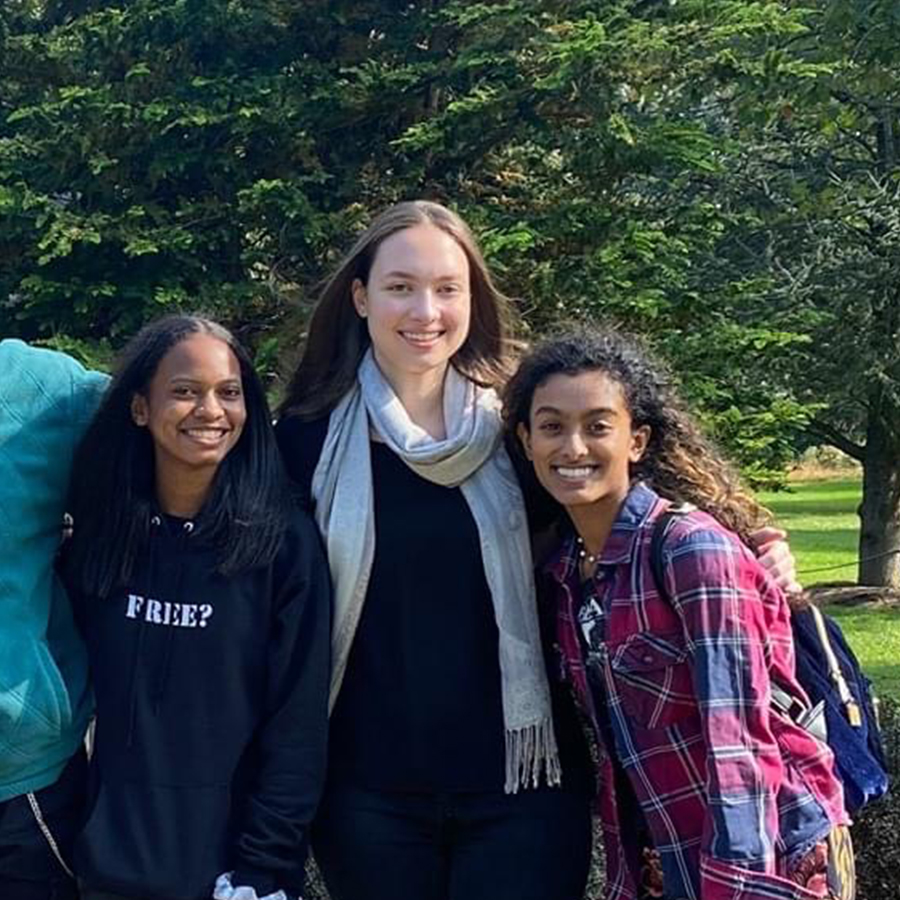 Three people smile at the camera as part of a larger group photo in a park.