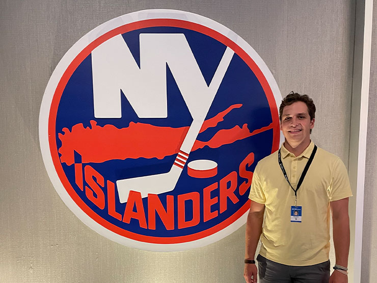 A college student in a yellow polo shirt stands with a lanyard around the neck next to the logo of the New York Islanders hockey team.