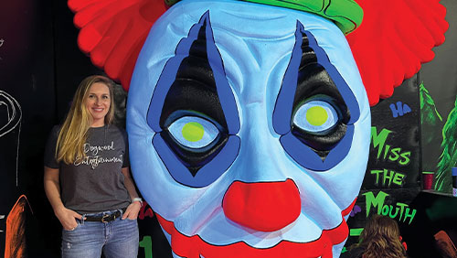 A woman stands next to a large, colorful and spooky clown prop at a mini golf course.
