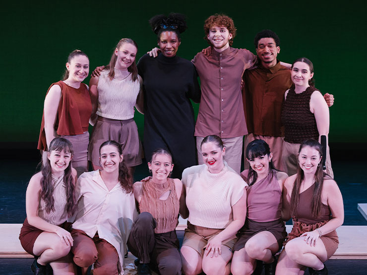 A group of students dressed in a variety of beige and brown tones pose together with an instructor, dressed in all black.