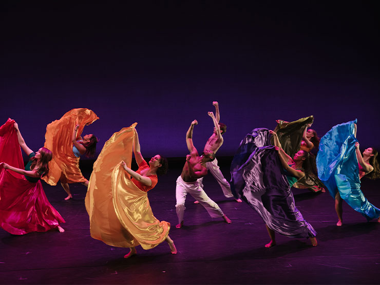 Students wearing colorful dresses dance expressively on a dark stage.