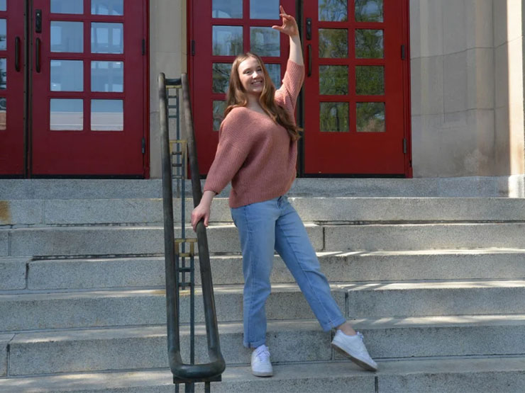 A college student strikes a dance pose on the steps to a campus building.