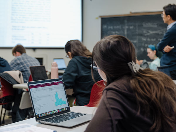 College students in a classroom sit, with open laptops displaying graphs, and look to the front of a classroom.